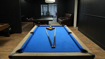 Une table de billard avec deux queues et des billes, un fauteuil en cuir et un banc en bois, dans une pièce aux murs sombres et dont la porte est visible.