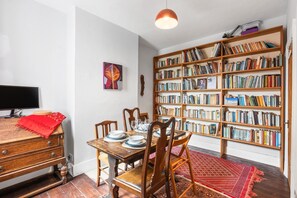 A dining area with a wooden table, chairs, a red rug, a bookshelf filled with books, and a television on a wooden dresser. - Daphne House By My Getaways (Brighton)