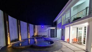 A modern outdoor pool area with a curved design, a covered seating area, and a two-story building with balconies. - Cozzy Villa Rans Cimacan (cianjur)