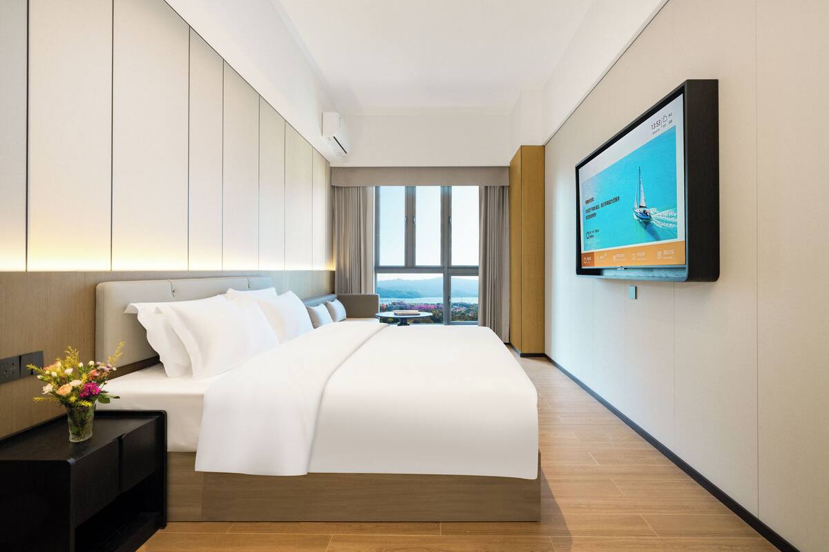 A hotel room with a bed, a TV mounted on the wall, a window with a view, and a small table with a vase of flowers.