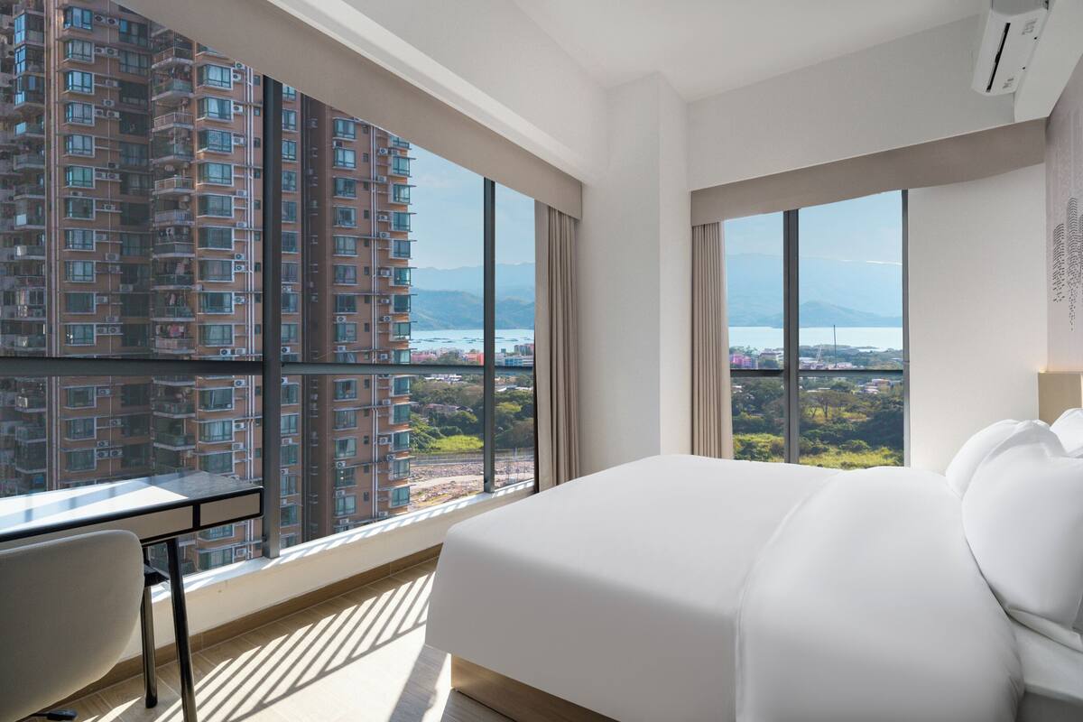 A hotel room with a large bed, a desk, and a view of a high-rise building and landscape.