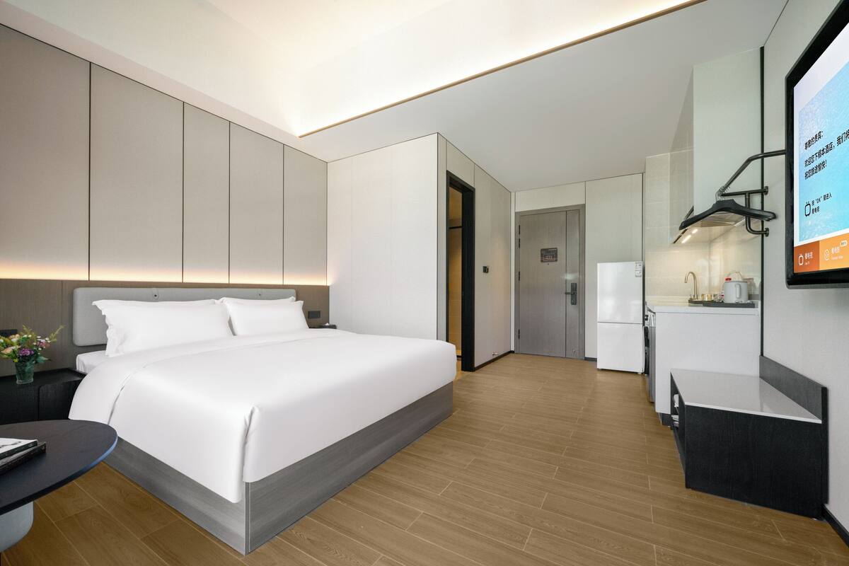 A modern hotel room with a large bed, a desk, a small kitchenette, and a television.