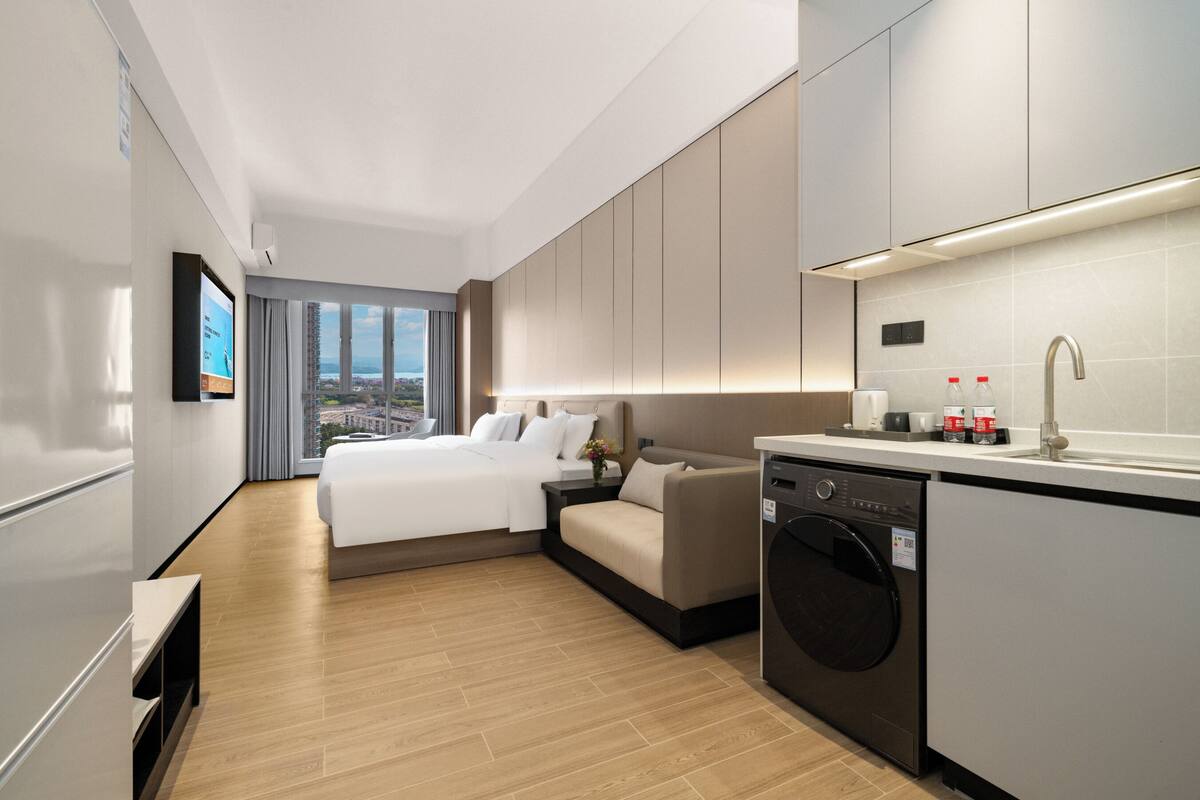 A modern hotel room with a bed, sofa, washing machine, and a view of the city.