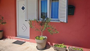Ein Gebäude mit roter Fassade, einer weißen Tür mit einem rautenförmigen Fenster und einem Fenster mit blauen Vorhängen. Vor der Tür steht ein Blumentopf.