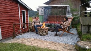 Two people sitting by a fire pit in an outdoor setting with a red wooden building and a vehicle in the background.