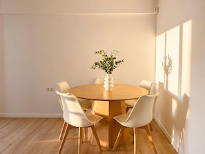 Ein runder Holztisch mit sechs weißen Stühlen, eine weiße Vase mit Grünpflanzen und eine wandmontierte Leuchte.