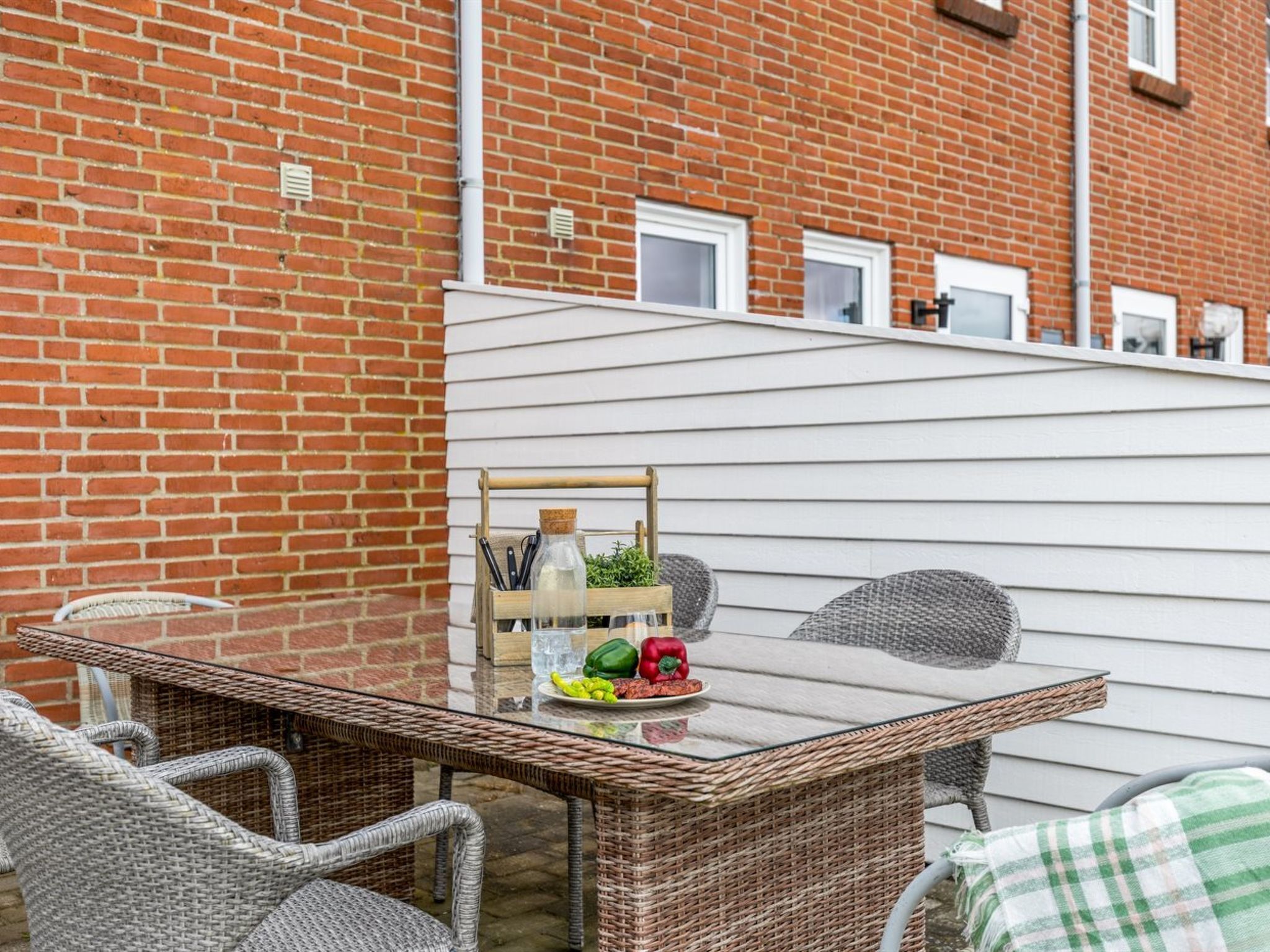 A glass-top table with wicker chairs, a small wooden side table with kitchen utensils, and a view of a brick building with windows.