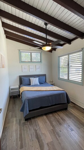 A bedroom with a bed, a ceiling fan, a window with blinds, and a small bedside table. - Yarianna’s Beach Apt. 2 (Aguadilla)