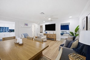 A modern living room with a wooden dining table, a sofa, a TV, and a kitchen in the background. - Steps to Beach | Family Retreat w/ Community Pool (Indian Shores)