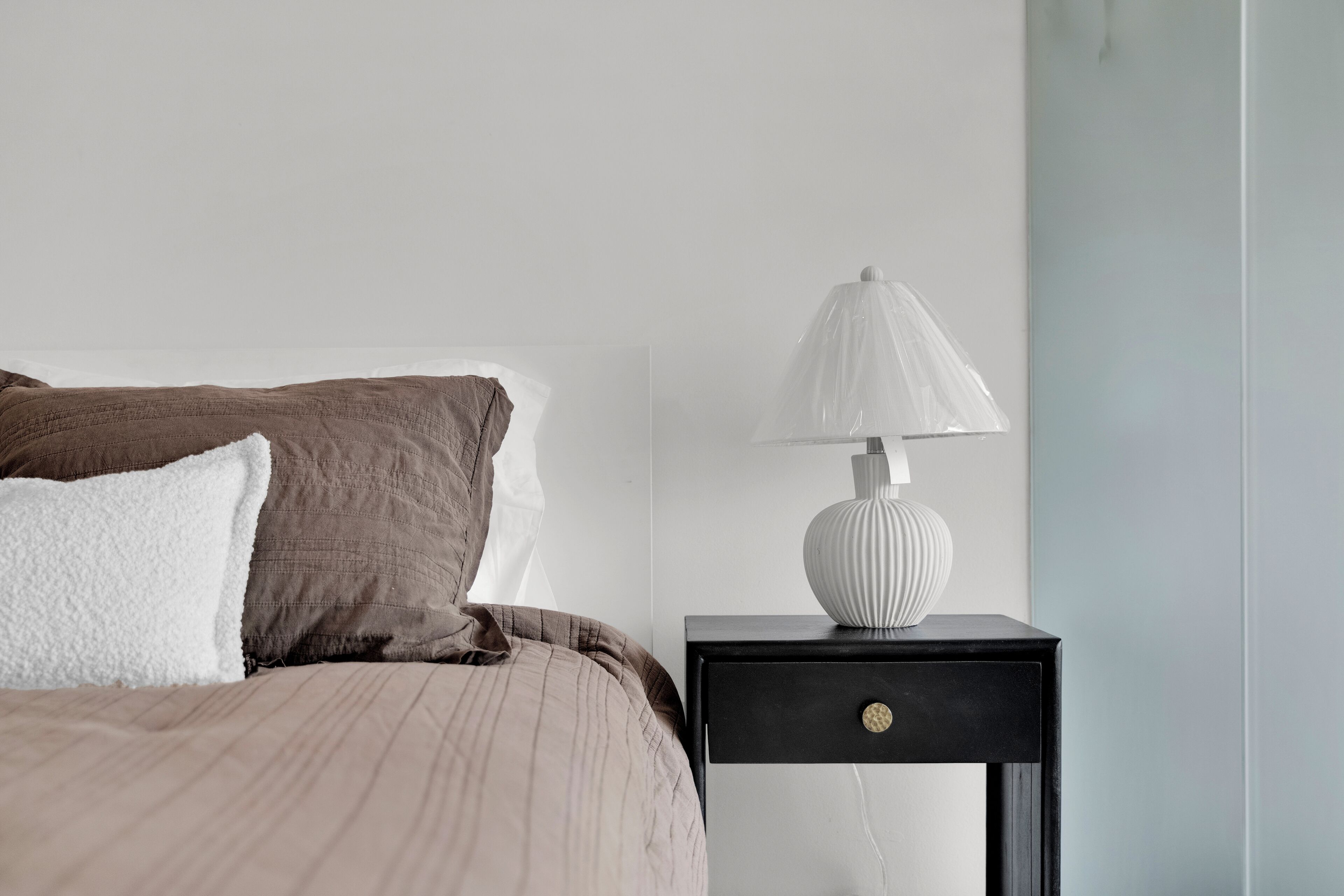 A neatly made bed with a striped bedspread and two pillows, a white lamp on a black nightstand, and a plain wall.