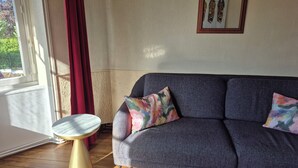 Een kamer met een grijze bank, een klein rond tafeltje, een spiegel aan de muur en een raam met gordijnen.