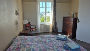 Een slaapkamer met een bloemenbedekker, een houten dressoir, een stoel, een raam met gordijnen en een radiator.