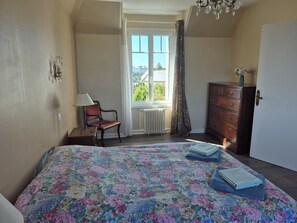 Ein Schlafzimmer mit floralem Bettbezug, einem Holzkasten, einem Stuhl, einem Fenster mit Vorhängen und einer Heizung.