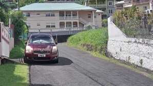 Une voiture de couleur bordeaux est garée sur une route étroite, avec un bâtiment en arrière-plan.