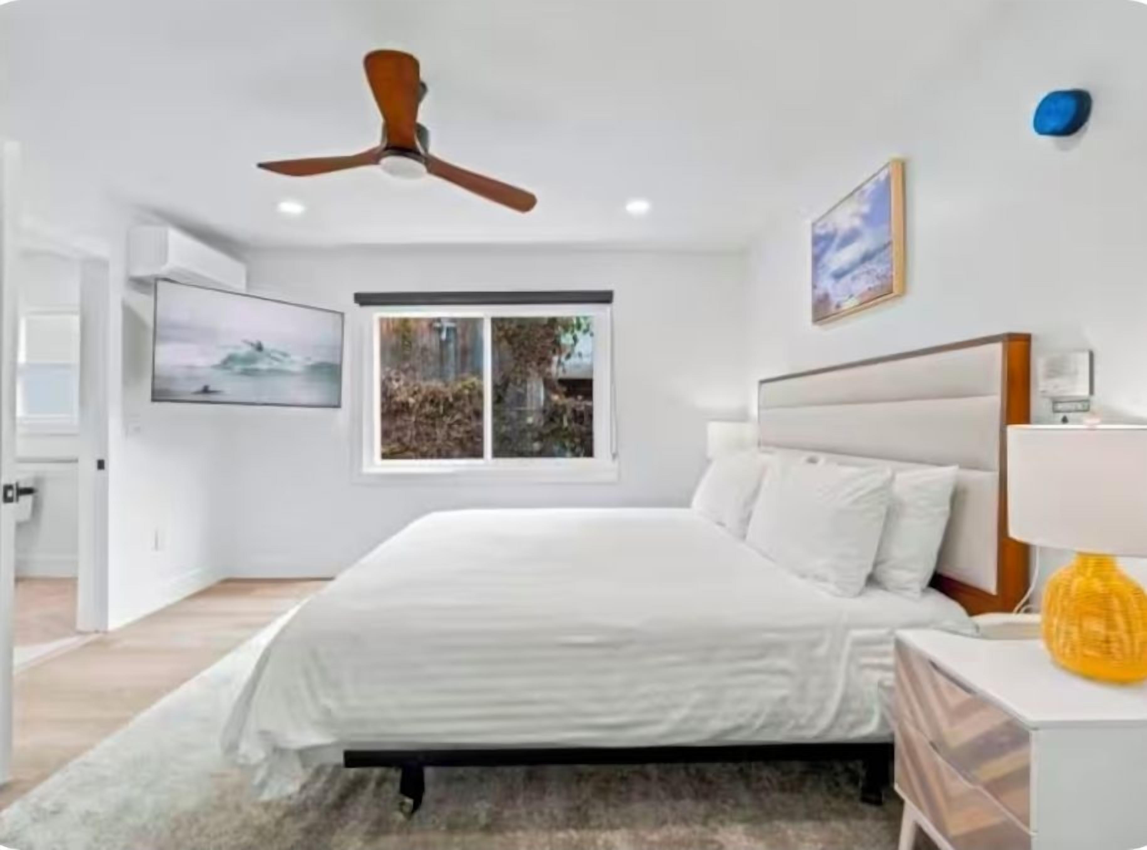 Une chambre à coucher avec un lit, un ventilateur de plafond, une fenêtre, une table de chevet avec une lampe et un tableau accroché au mur.