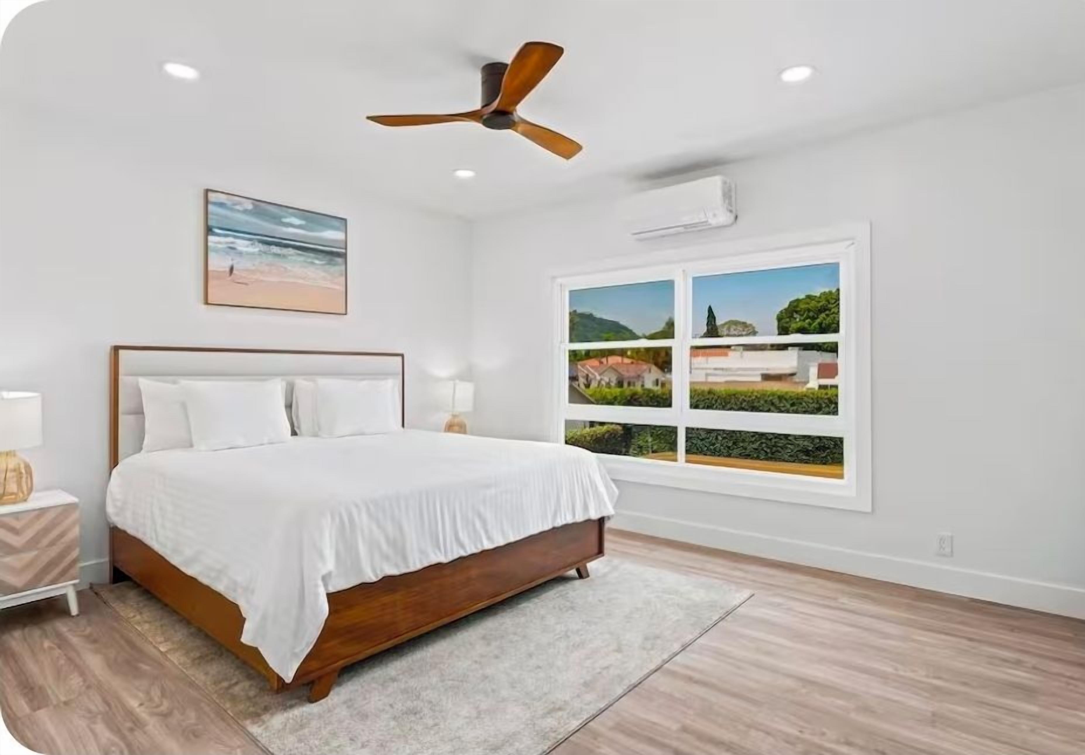 A bedroom with a bed, a ceiling fan, a window with a view, and a nightstand with a lamp.