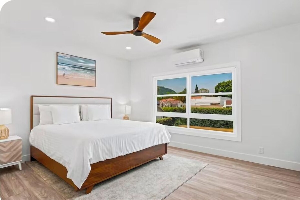 A bedroom with a bed, a ceiling fan, a window with a view, and a nightstand with a lamp.