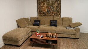 A living room with a brown sofa, a coffee table with chess and a toy car, and a tapestry on the wall. - Cozy 2 bedroom Apartment with Arcade & Games (Cincinnatus)