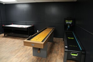 A billiards table, a shuffleboard table, and a billiards score machine in a room with dark walls and wooden flooring.