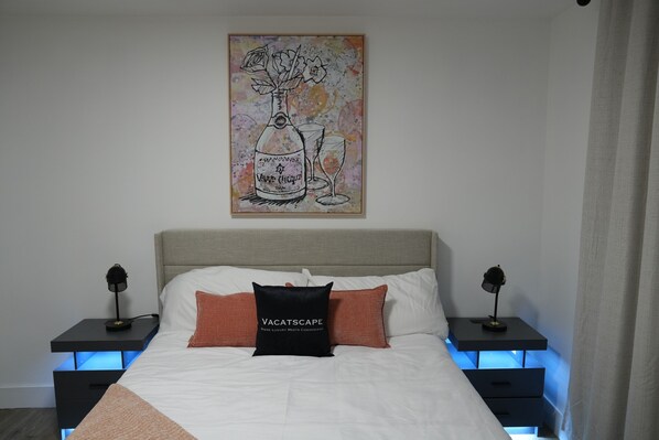 A bedroom with a bed, two bedside tables with lamps, a wall-mounted artwork, and a nightstand with drawers.