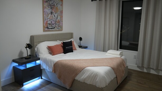 A modern bedroom with a bed, bedside tables, a night lamp, a window with curtains, and a wall art piece. - Cozy 2 bedroom Apartment with Arcade & Games (Cincinnatus)