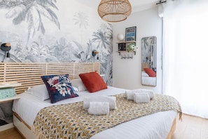 Ein Schlafzimmer mit einem Bett, einem Kopfteil, einem an der Wand befestigten Spiegel und tropisch gestalteter Tapete.