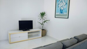 Una sala de estar con un televisor de pantalla plana sobre un mueble blanco, un sofá gris, una planta en maceta y un cuadro en la pared.