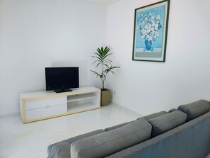 Uma sala de estar com uma TV de tela plana em um armário branco, um sofá cinza, um vaso de planta e um quadro na parede.