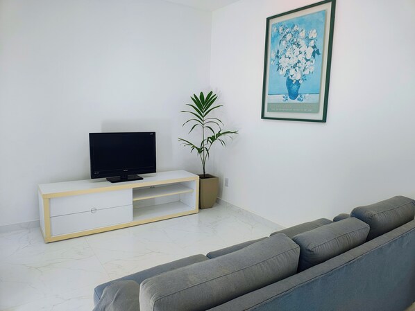 Un salon avec un téléviseur à écran plat posé sur un meuble blanc, un canapé gris, une plante en pot et un tableau encadré accroché au mur.