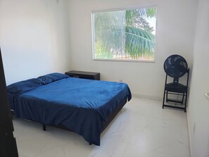 Une chambre à coucher comprenant un lit, une table de chevet, une chaise, un ventilateur sur pied et une fenêtre donnant sur un paysage verdoyant.