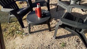 An outdoor seating area with two Adirondack chairs and a small round table, a red thermos, and a broom leaning against a wall.