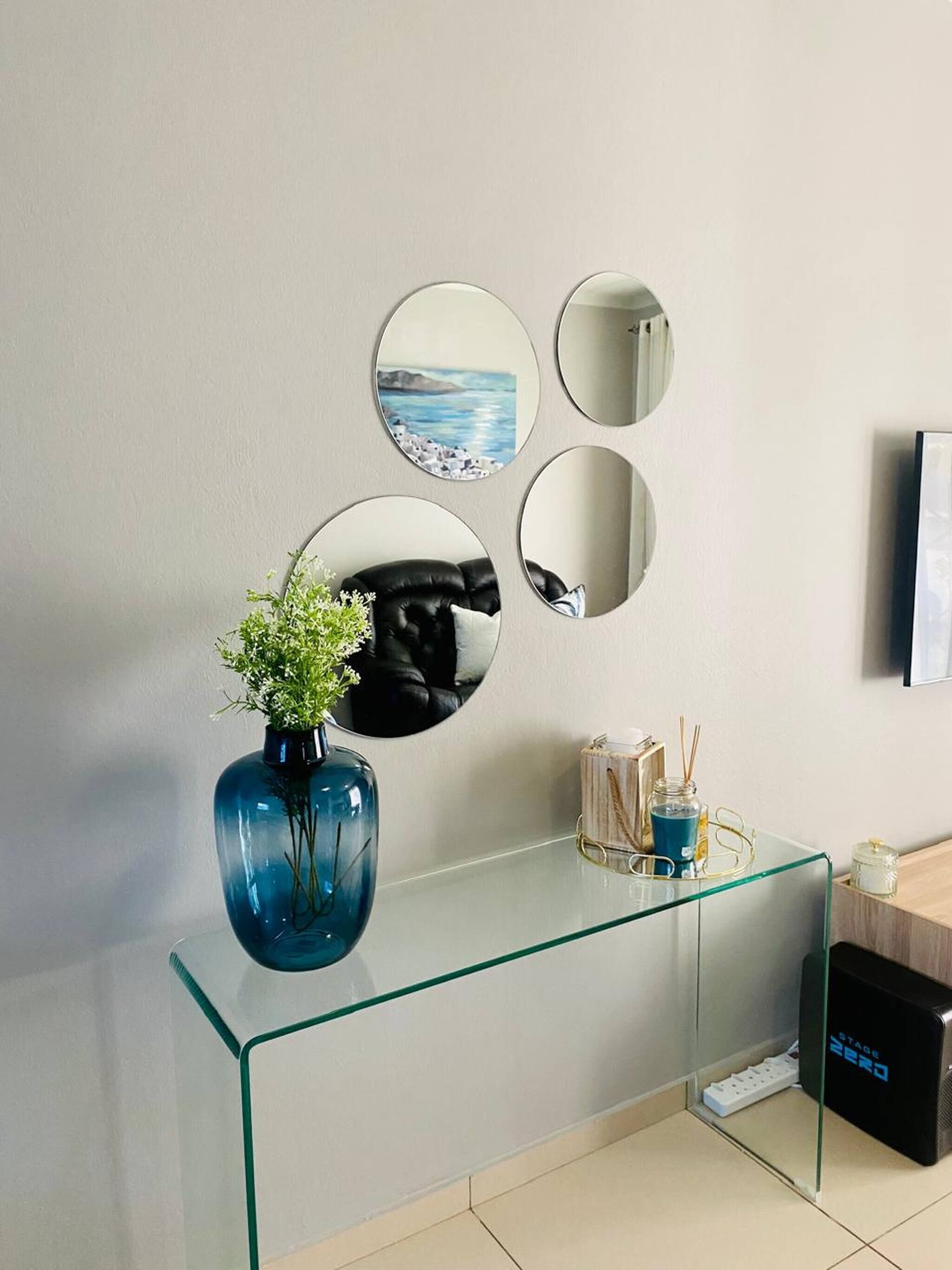 A modern bathroom with a glass vanity, a blue vase with greenery, and a mirror with a coastal painting.