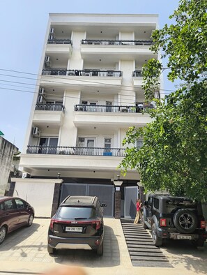 A multi-story building with balconies, a gated entrance, and parked vehicles. - SkyNest stays- A private terrace garden | Near cyber hub  & galleria market |  (Gurugram)