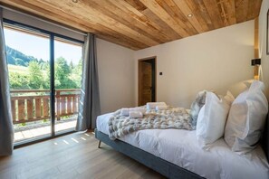 A bedroom with a large bed, a wooden ceiling, and a balcony with a view of greenery. - Chalet des Granges • 12 Guests • Spa & Private Cinema (Morzine)