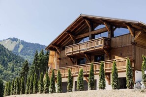 Un chalet en bois doté d’un balcon, entouré d’arbres à feuilles persistantes et avec des montagnes en arrière-plan.