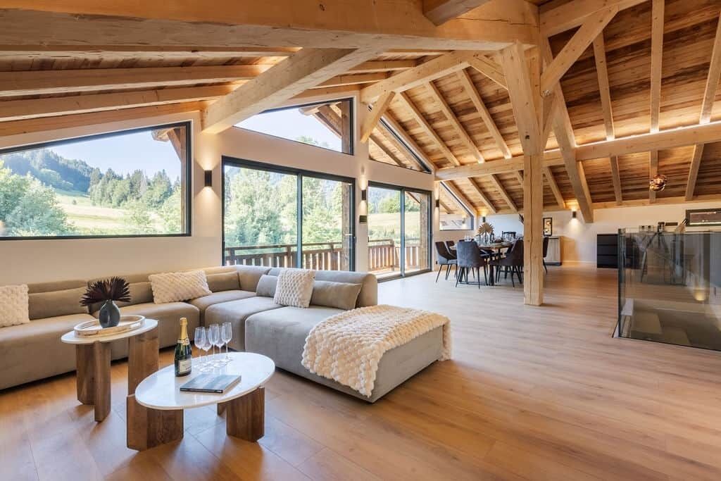 Een ruime woonkamer met houten plafond, grote ramen, een bank, een salontafel en een eethoek.