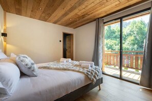 Ein Schlafzimmer mit einem Bett, einer Holzbalkendecke, einem Balkon mit Blick auf Bäume und einer Tür zu einem anderen Raum.
