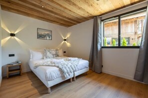 Ein Schlafzimmer mit Holzboden, einem Bett mit weißer Bettwäsche, zwei Wandleuchten, einem Fenster mit Vorhängen und einem kleinen Nachttisch.