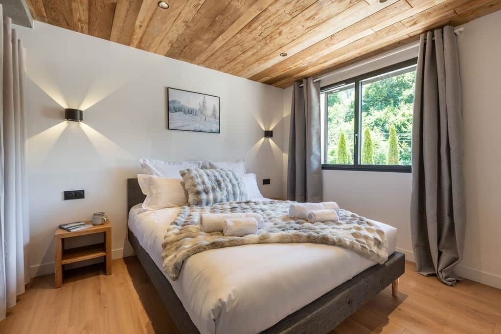 A bedroom with a wooden ceiling, a bed with a white comforter, two wall-mounted lights, a small bedside table, and a window with curtains.