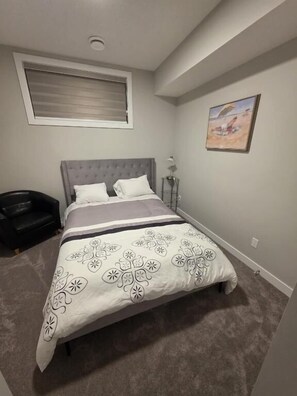 A bedroom with a bed, a nightstand, a lamp, a window with blinds, and a painting on the wall. - Spacious and Modern 2-bedroom Apartment in Calgary (Calgary)
