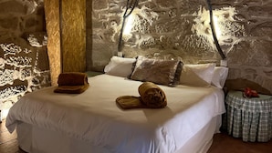 Un dormitorio con pared de piedra, cama con ropa blanca, cabecera de madera y una mesita con teléfono.