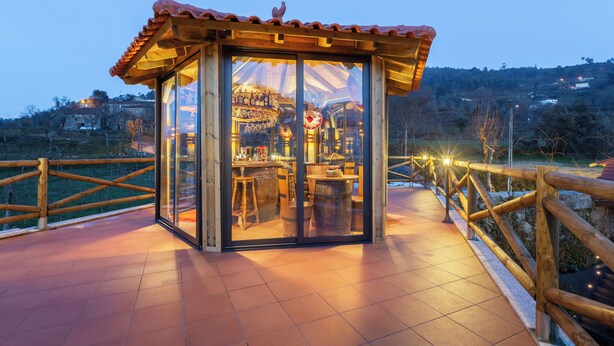 Una estructura con paredes de vidrio, techo de madera, cercado de madera y vista a un área de bar con mobiliario de madera.