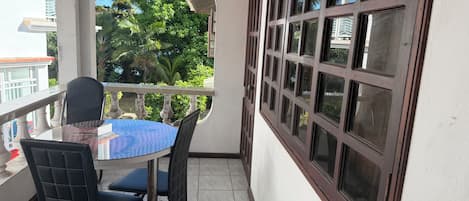 A balcony with a round table and chairs, a view of greenery, and a building in the background.