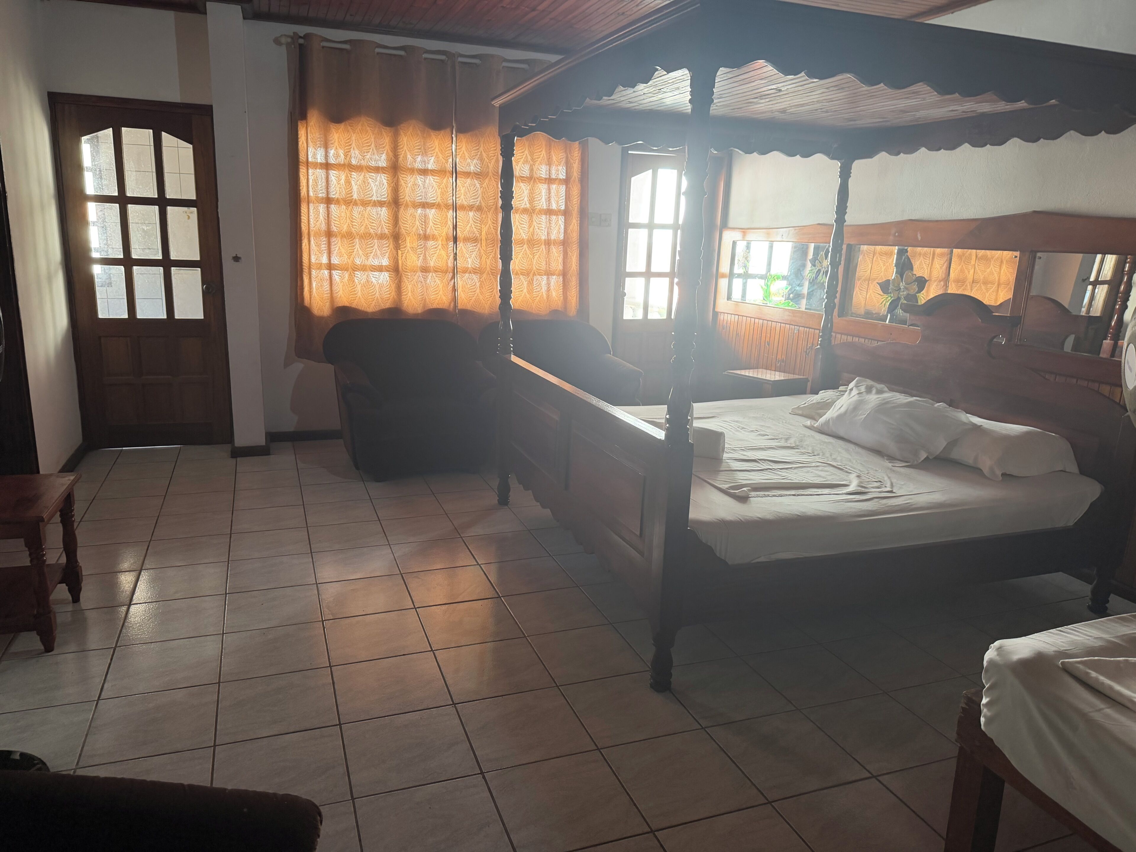 A room with a four-poster bed, a wooden chair, a sofa, and a tiled floor.