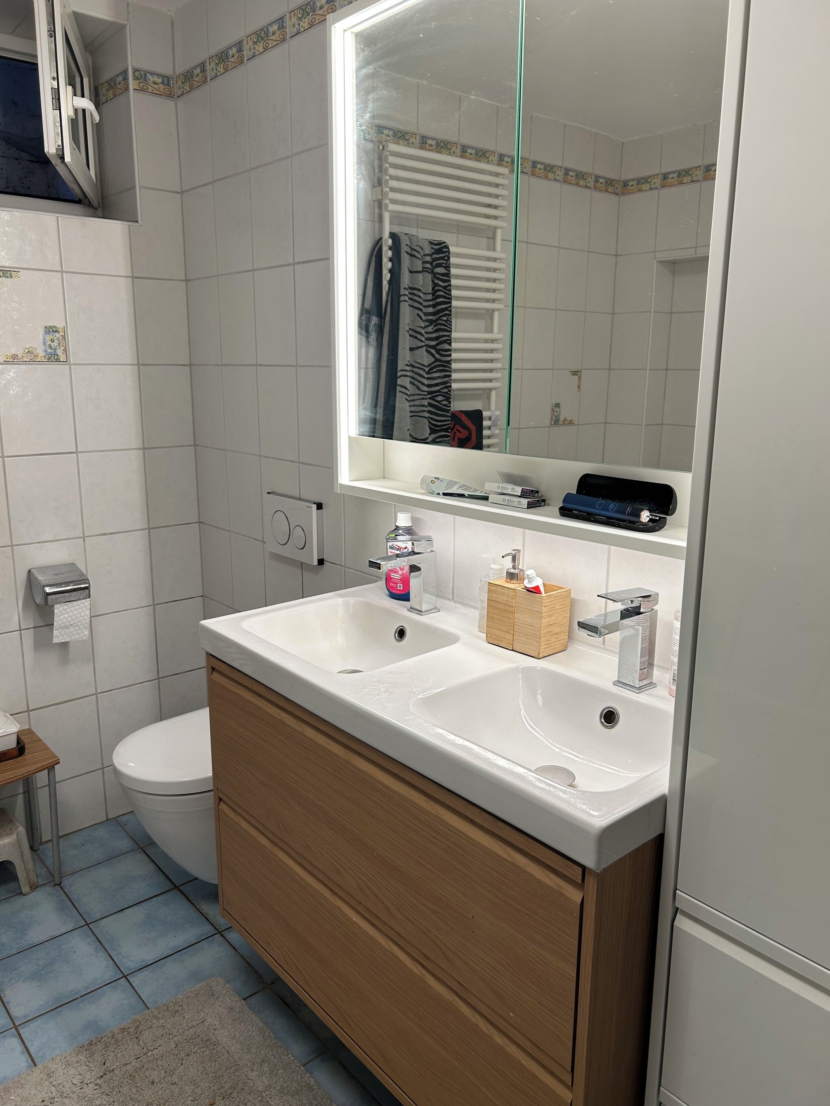Un baño con lavamanos blanco, tocador de madera, un espejo, inodoro y perchero para toallas.