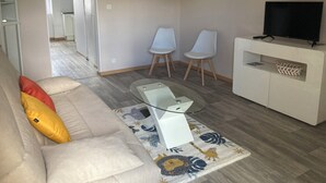 Ein Wohnzimmer mit einer Couch, zwei Sesseln, einem Glastisch, einem Fernseher auf einem Schrank und einem gemusterten Teppich.