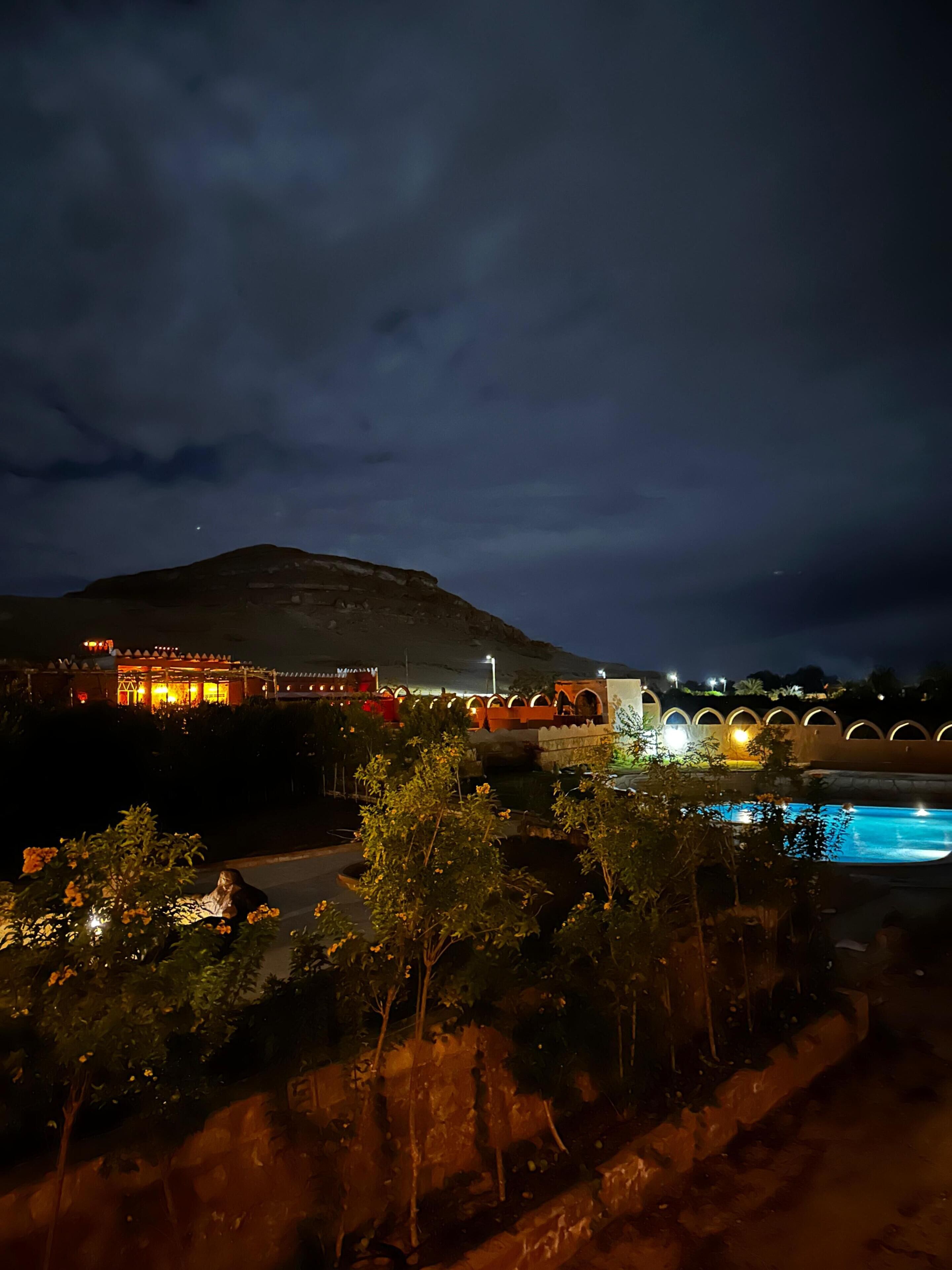 Vista nocturna de un hotel con piscina, una montaña al fondo y un edificio iluminado con arcos.