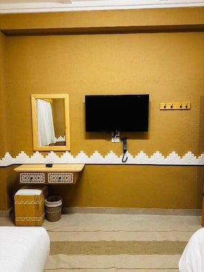 A hotel room with a bed, a television mounted on the wall, a mirror, a small wooden cabinet, and a trash bin. - Happy Oasis Hotel (Siwa)