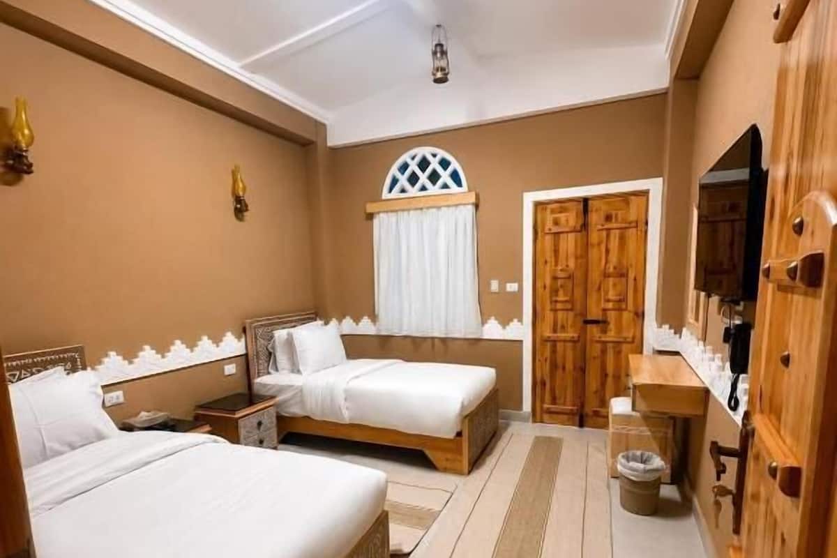 A hotel room with two beds, a wooden wardrobe, a small table, and a window with curtains.