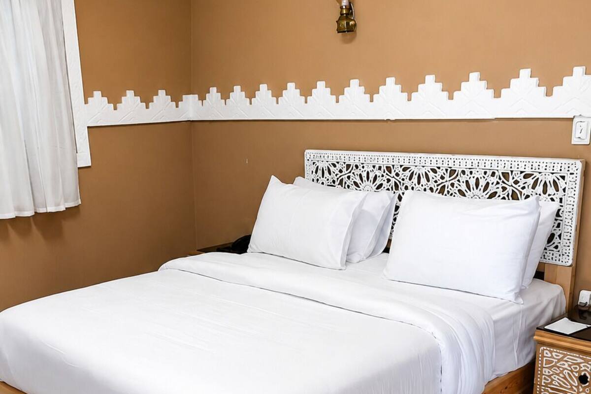 A bed with white bedding, a wooden headboard, a nightstand with a patterned tray, and an air conditioning unit on the wall.